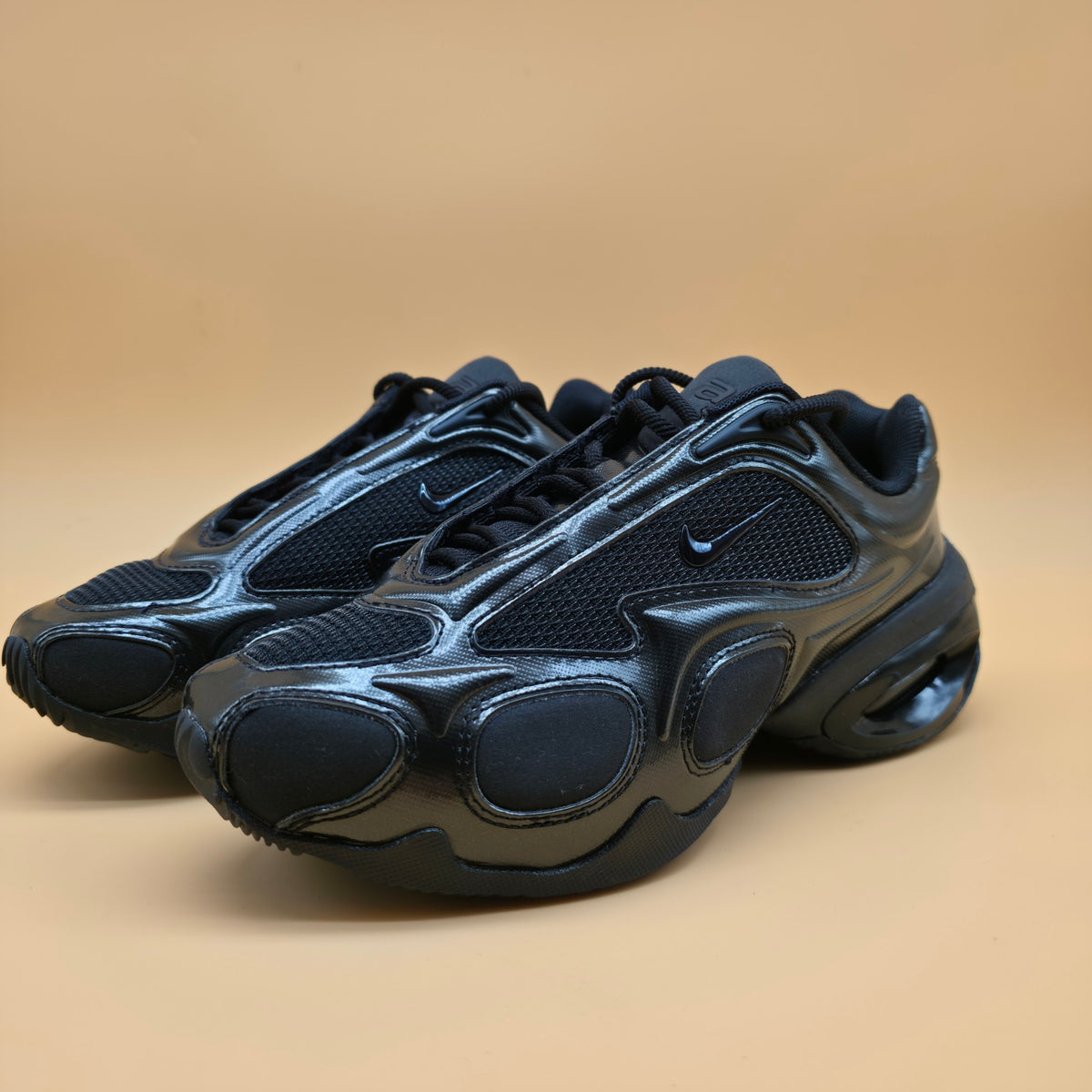 A pair of Nike Air Max 2013 sneakers in black, featuring a glossy synthetic leather overlay, mesh panels, and a thick cushioned sole, set against a solid beige background.