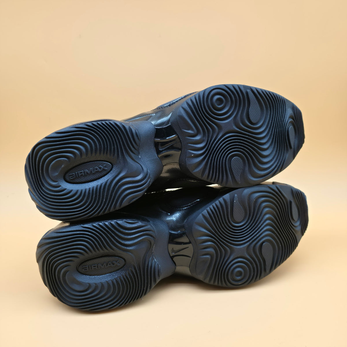 A pair of dark blue athletic shoes shown from the sole view, highlighting their textured, wavy tread pattern and 'AIRMAX' branding, set against a solid beige background.