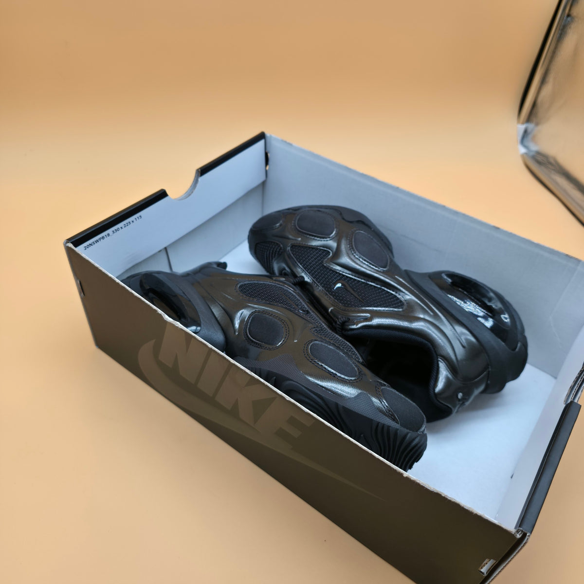 A pair of black Nike sneakers inside an open black Nike shoebox, photographed on a warm beige studio background.