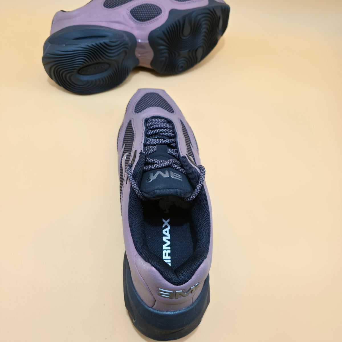 A pair of athletic sneakers shown on a plain beige studio background, with one shoe standing upright to display the side and heel and the other placed upside down to show the sole.