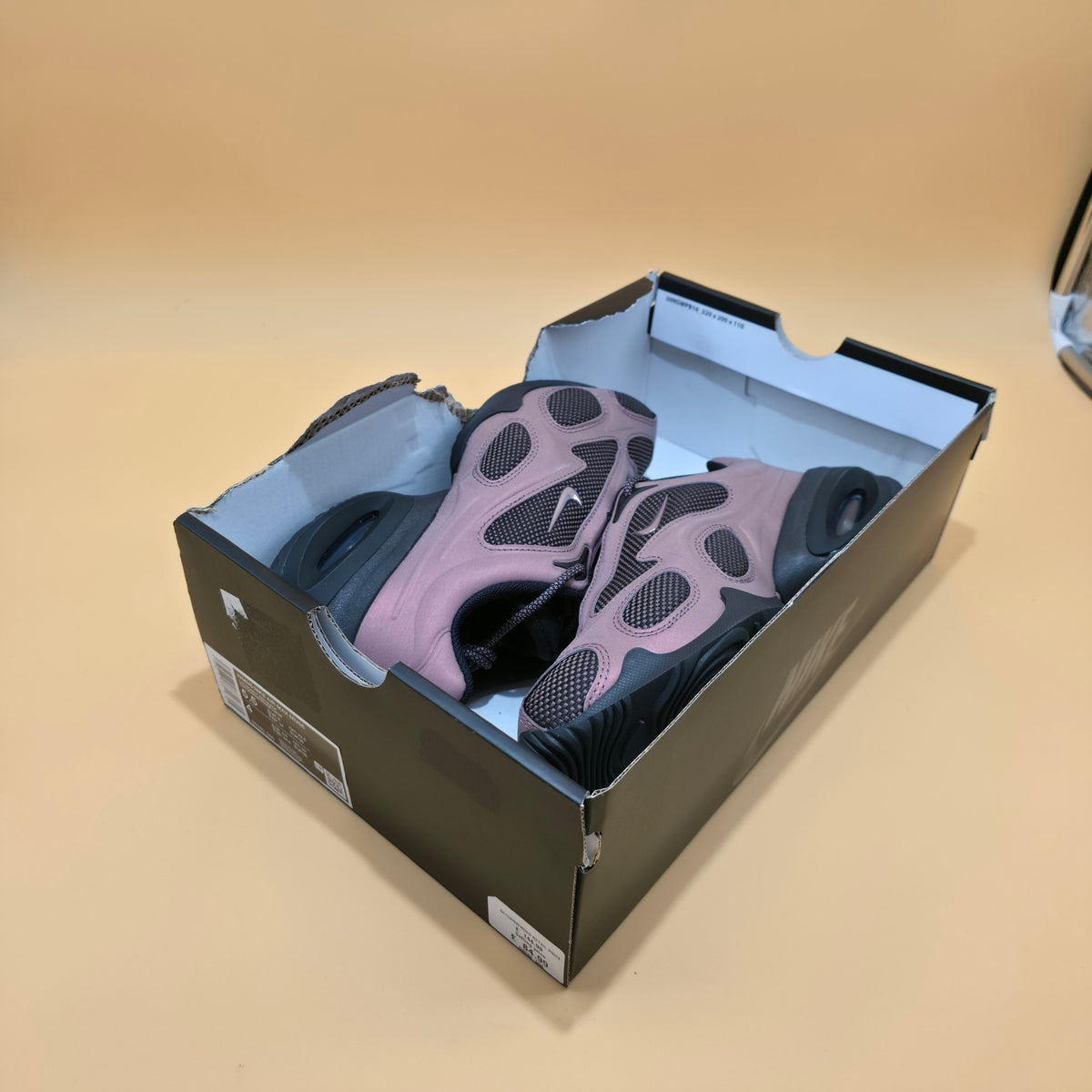 A pair of Nike running shoes in dusty pink and black, presented inside their original dark gray Nike shoebox on a solid beige background.