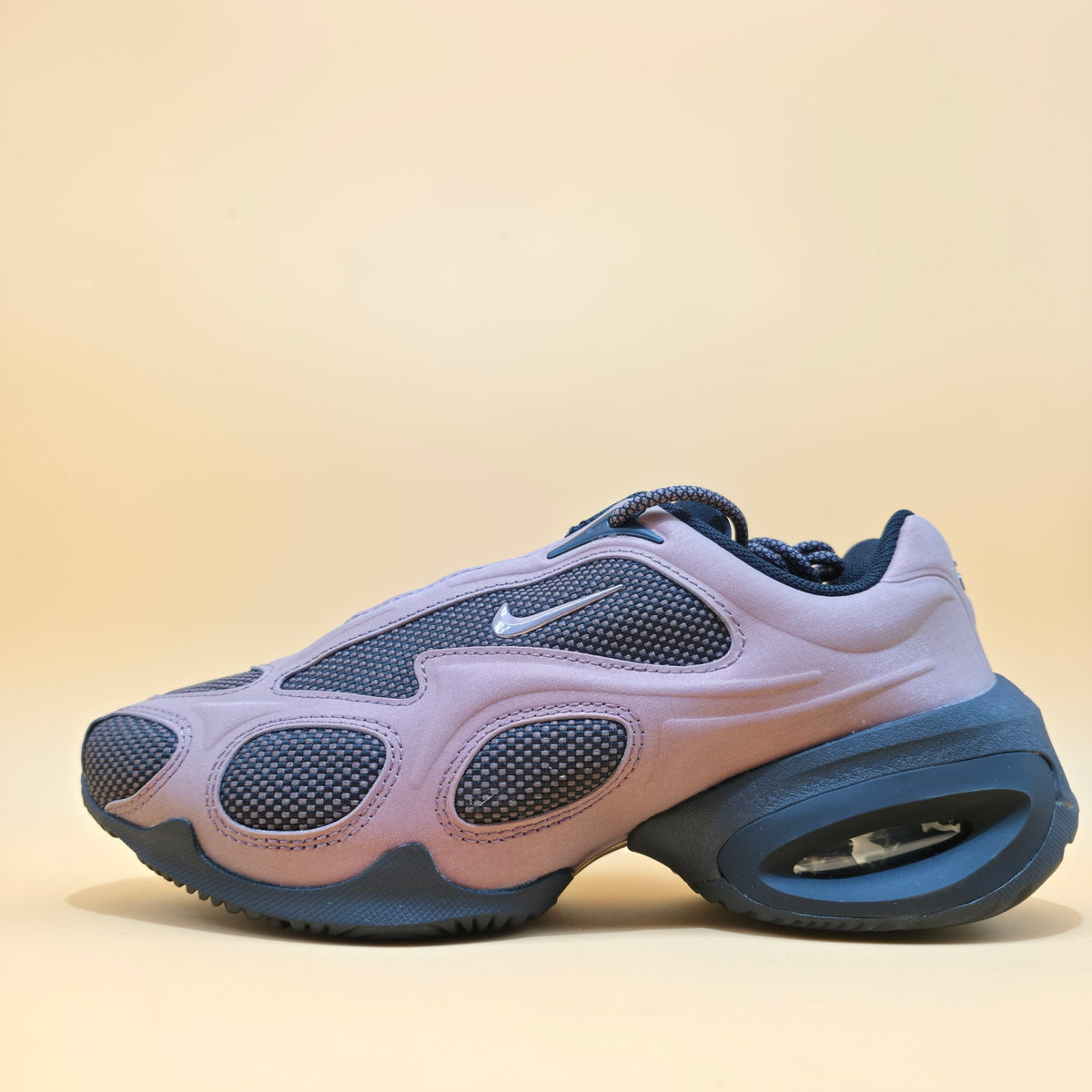 A Nike sneaker in a dusty lavender and navy blue colorway, featuring a thick textured sole and a visible white Swoosh logo on the side, set against a solid beige background.