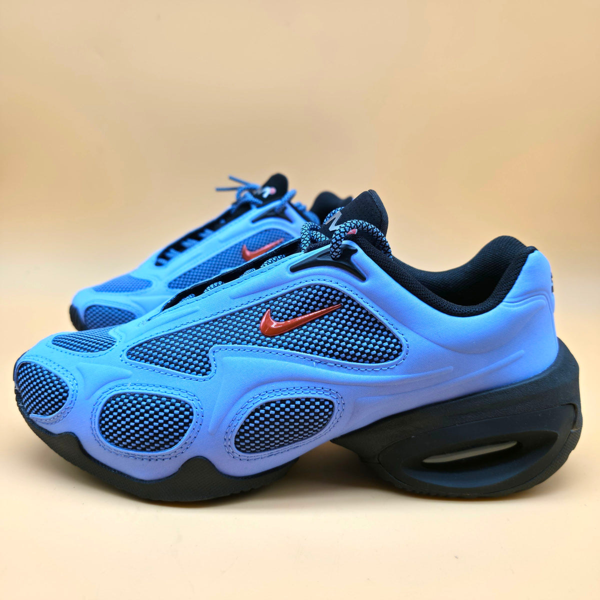A pair of Nike Air Max 90 sneakers in light blue and navy blue, featuring a signature orange Swoosh logo, set against a solid beige background.