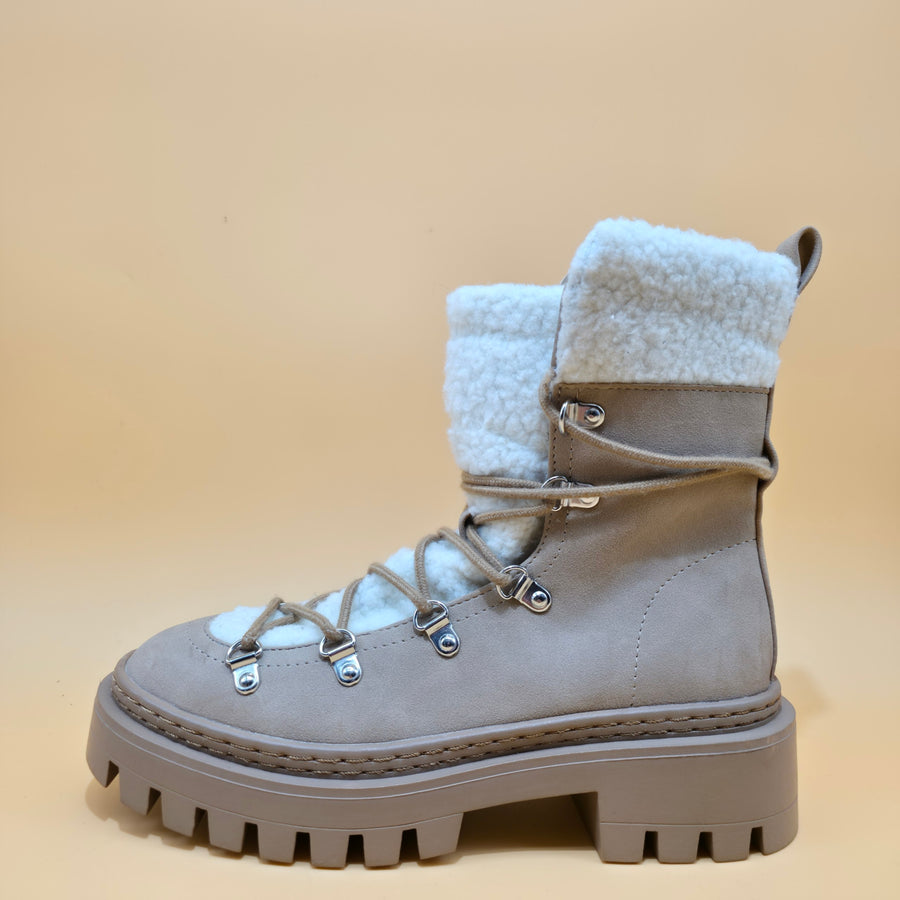 A light taupe suede winter boot with a thick lug sole, lace-up front, and a fluffy white shearling-style collar, shown against a solid beige background.