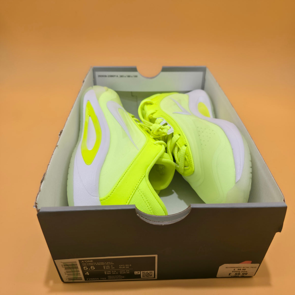 A pair of Nike sneakers in neon yellow-green and white, presented inside their original dark gray Nike shoebox against a solid orange background.