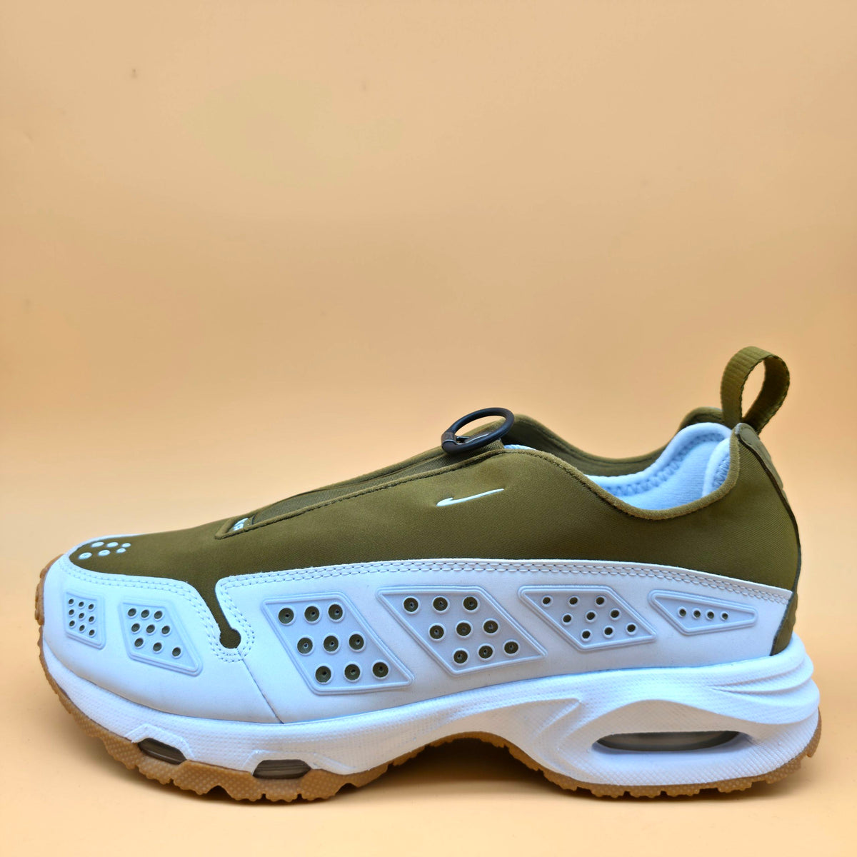 A Nike sneaker in olive green and white, featuring a slip-on design with a pull tab, geometric perforated side panels, and a chunky white midsole with a gum outsole, set against a solid beige background.