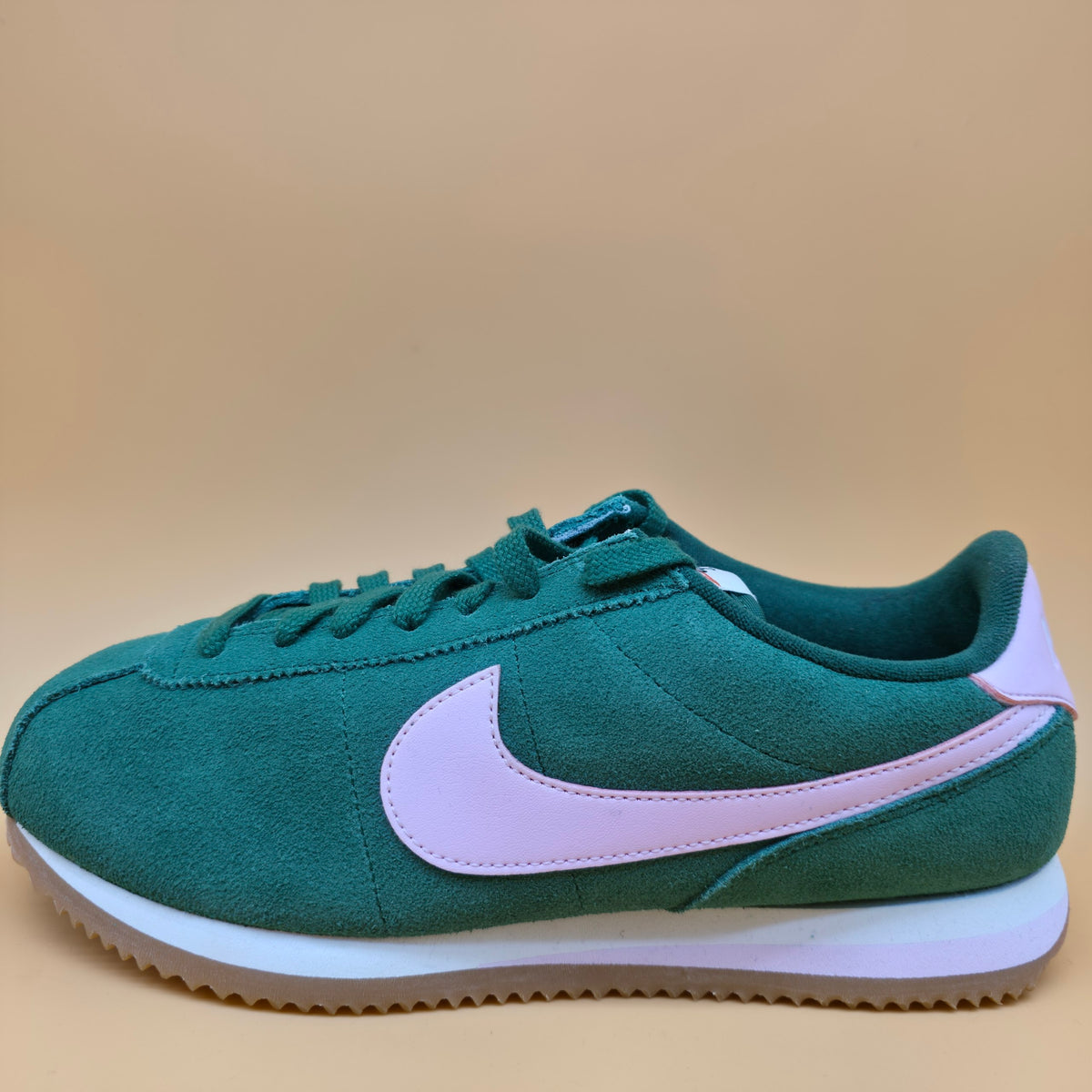 A Nike Classic Cortez sneaker in dark green suede with a light pink Swoosh and heel tab, featuring a white midsole and gum sole, set against a solid beige background.
