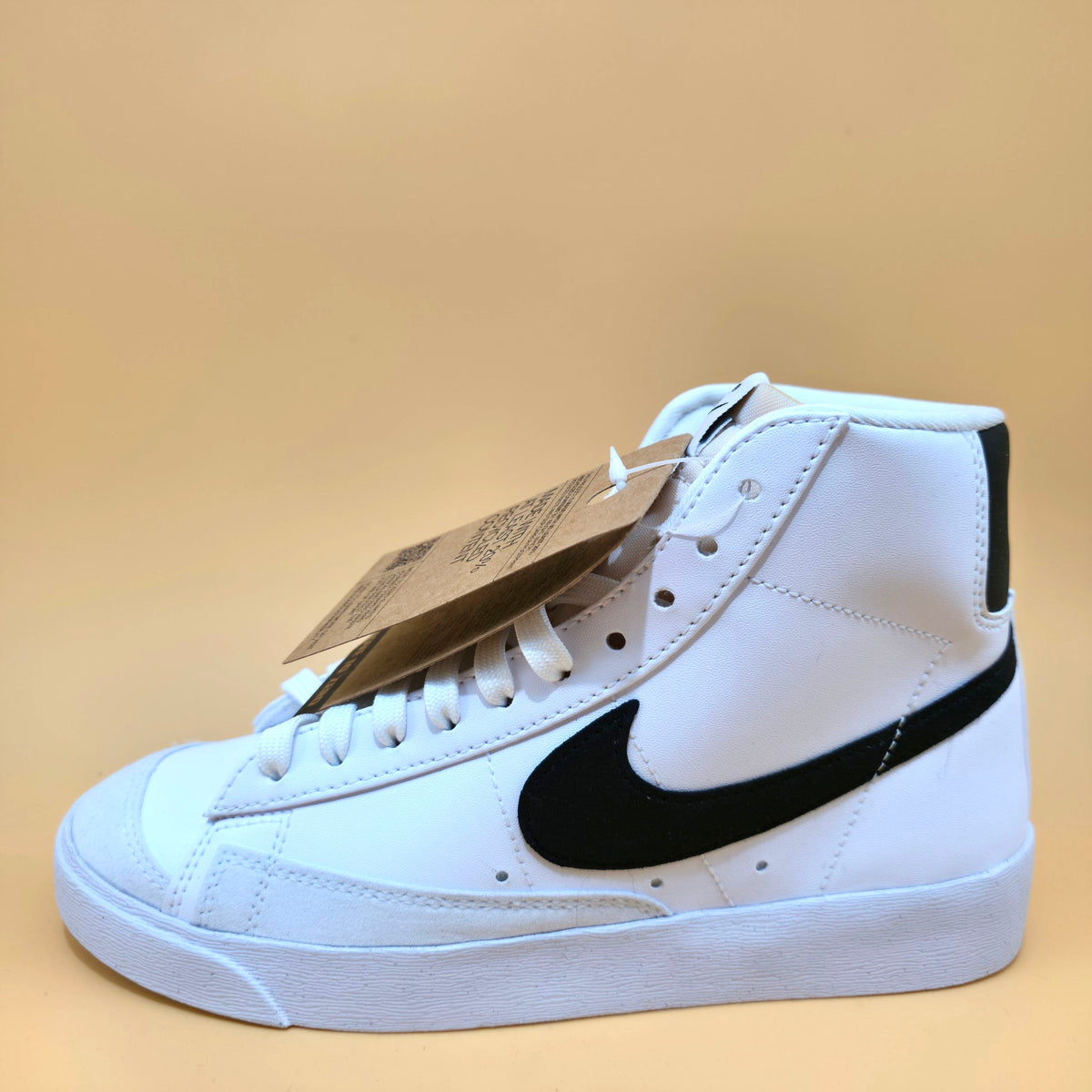 A white high-top sneaker with a black Nike Swoosh, shown from the side profile on a solid beige background. A brown paper tag is attached to the laces.