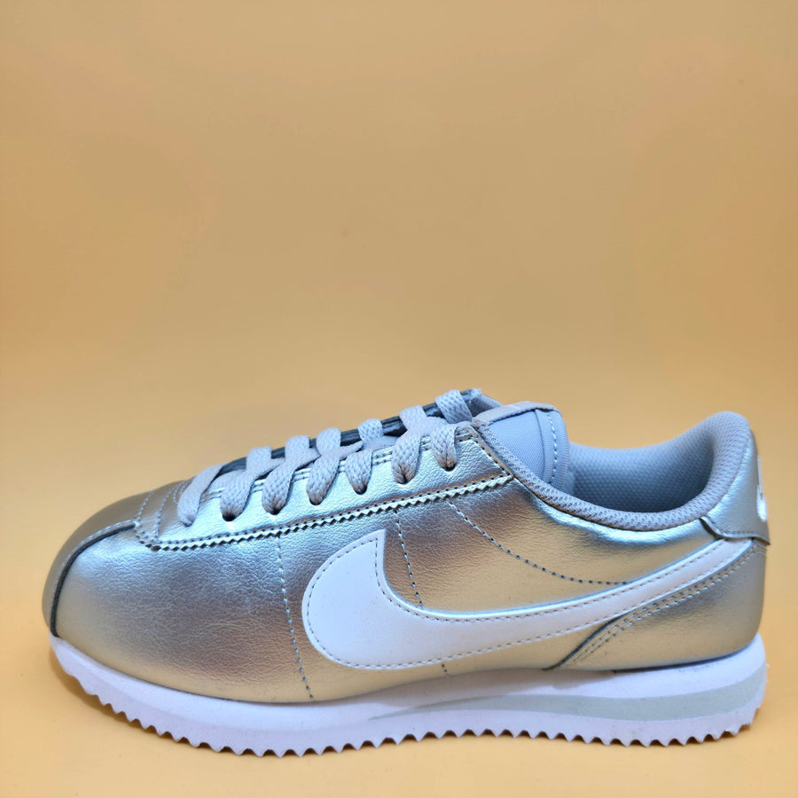 A metallic silver Nike Cortez sneaker with a white Swoosh and white sole, photographed from the side against a solid tan background.