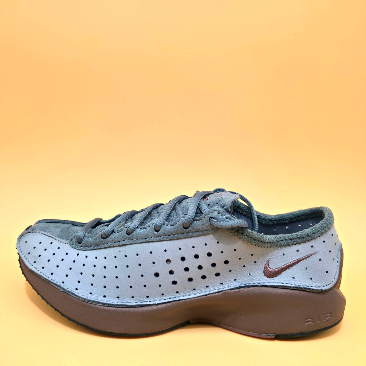 A Nike Air Max sneaker in a muted blue-gray colorway, featuring a perforated synthetic upper, suede overlays, matching gray laces, and a thick brown Air Max sole, set against a solid yellow-orange background.