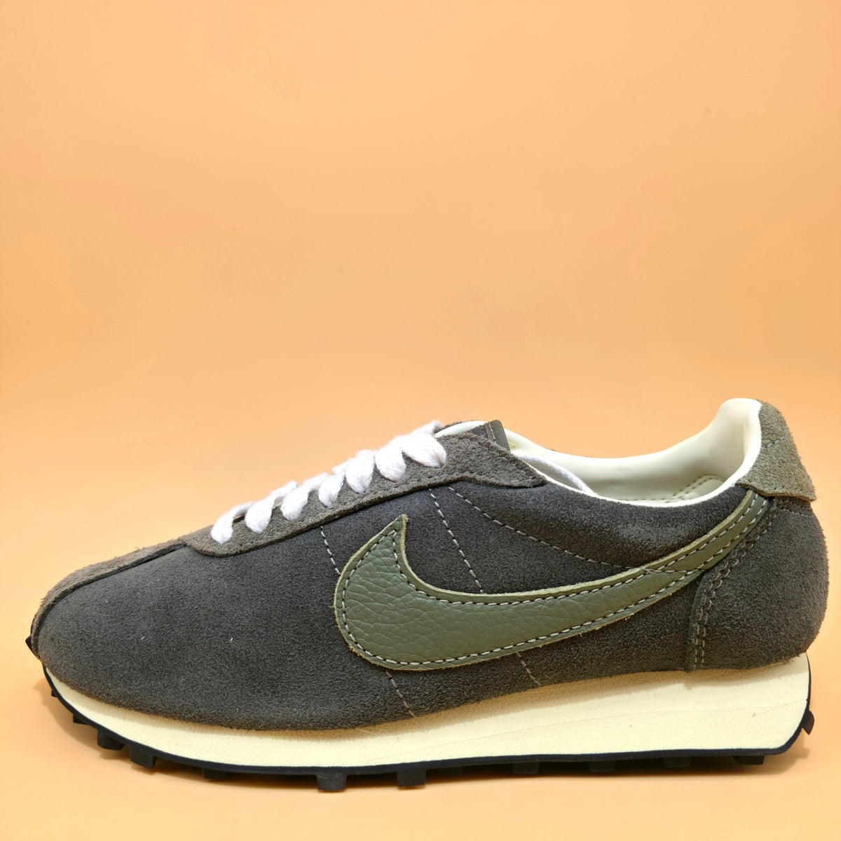 A Nike sneaker in dark gray suede and olive green leather, featuring white laces and a cream-colored midsole, set against a solid peach background.