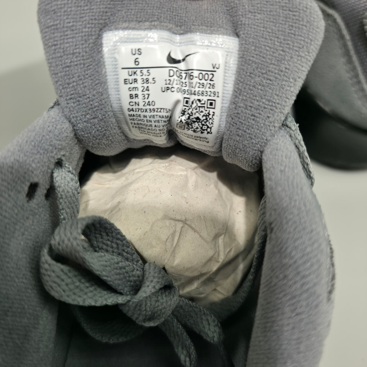 Close-up of the interior collar of a gray Nike sneaker, showing the size and model label with the Nike swoosh, along with the gray laces and white tissue paper inside the shoe.