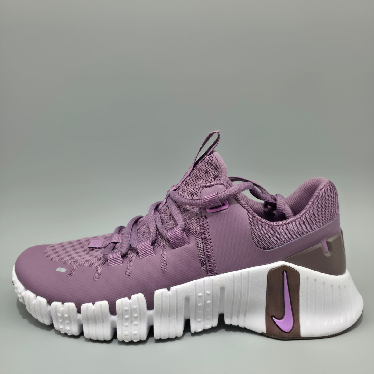 A Nike athletic sneaker in a dusty purple colorway, featuring a textured knit upper, matching purple laces, a white flexible sole, and a brown heel accent with a purple Nike Swoosh.