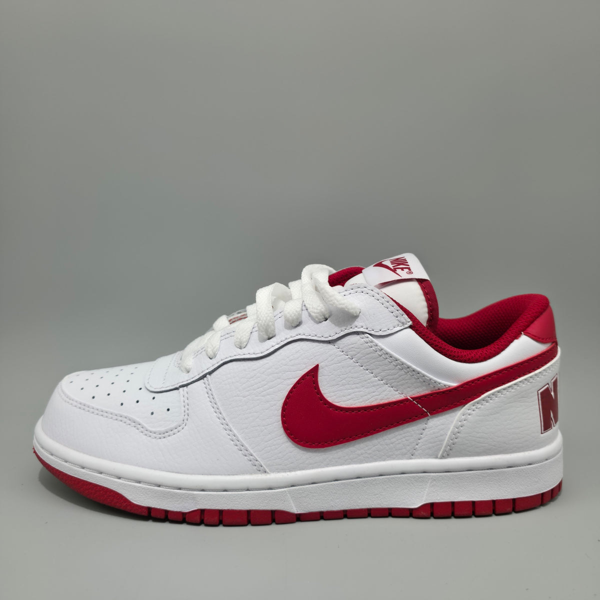 A white Nike Dunk Low sneaker with red accents, featuring a red Swoosh, red heel tab, and red outsole, shown from a side profile on a gray background.