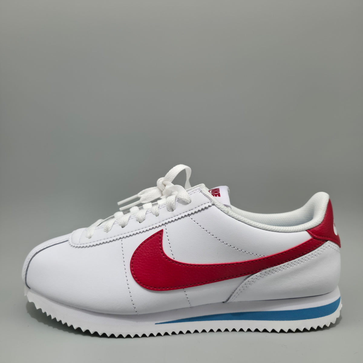 A white Nike Cortez sneaker with a red Swoosh, white laces, and a blue accent stripe on the sole, photographed against a neutral gray background.