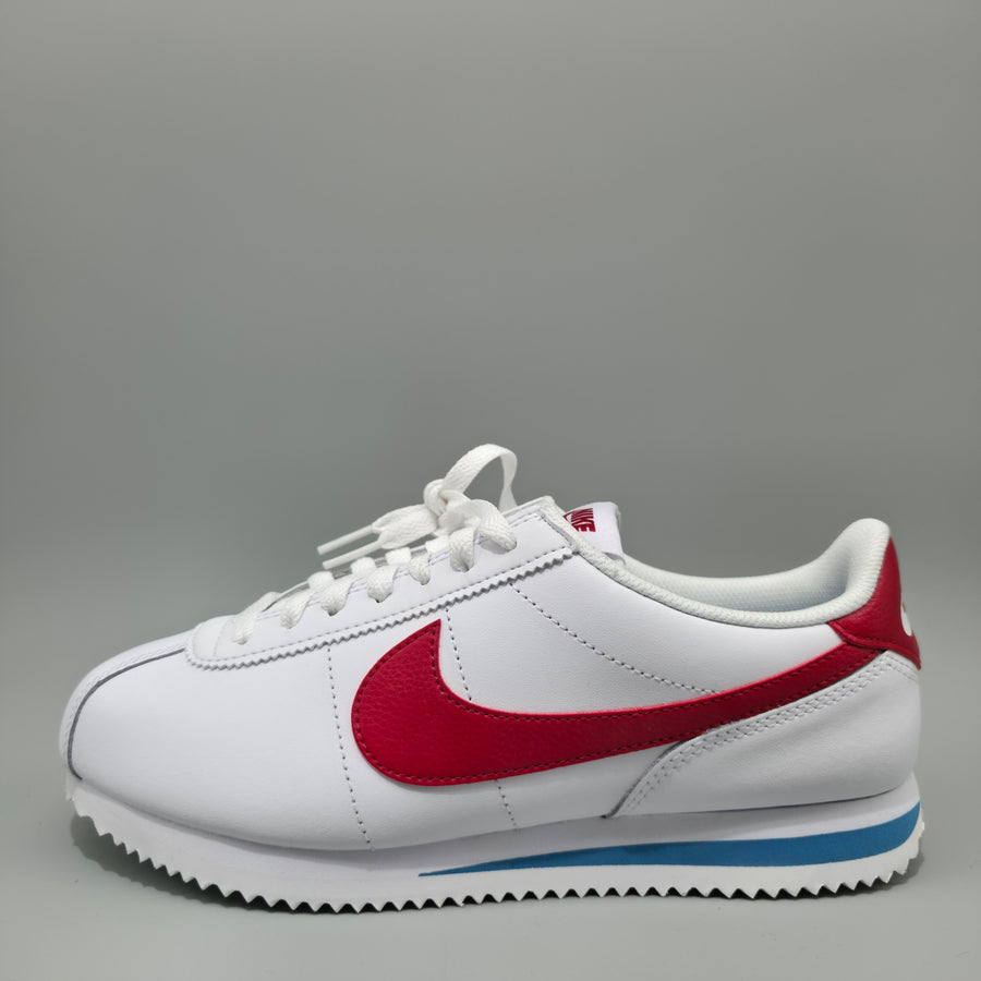 A white Nike Cortez sneaker with a red Swoosh, white laces, and a blue accent stripe on the sole, photographed against a neutral gray background.