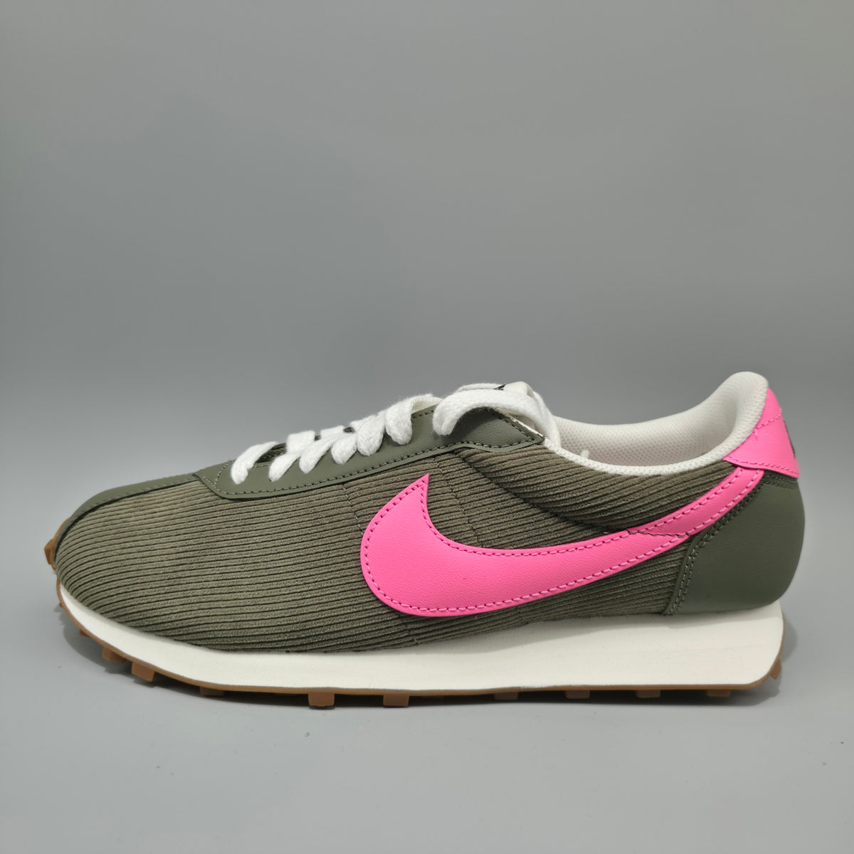 A Nike Classic Lifestyle sneaker in olive green corduroy with a vibrant pink Swoosh, white laces, and a white midsole with a gum outsole, shown against a neutral gray background.