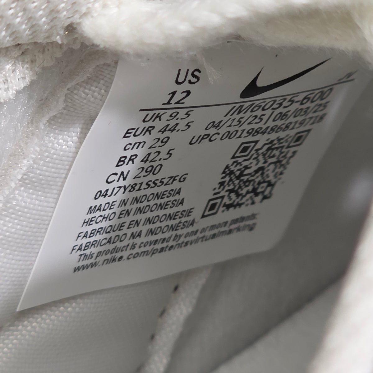 Close-up of a white fabric garment tag with a black Nike swoosh, size information, QR codes, and printed text on a white knit background.