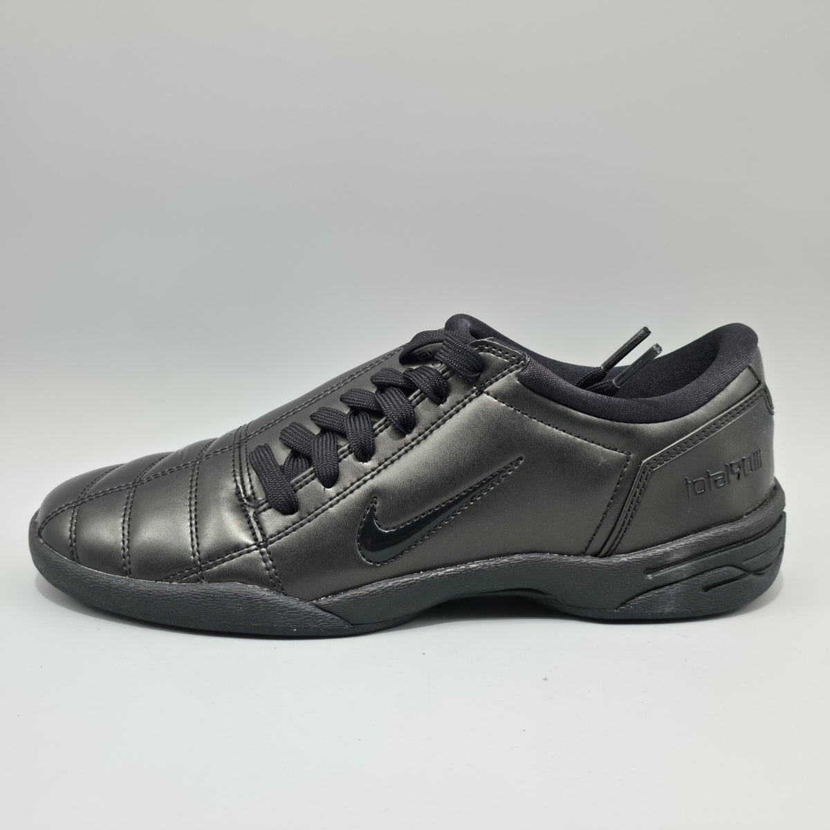 A black Nike indoor soccer shoe with a glossy leather upper, black laces, and a black sole, shown from a side profile view against a light gray background.