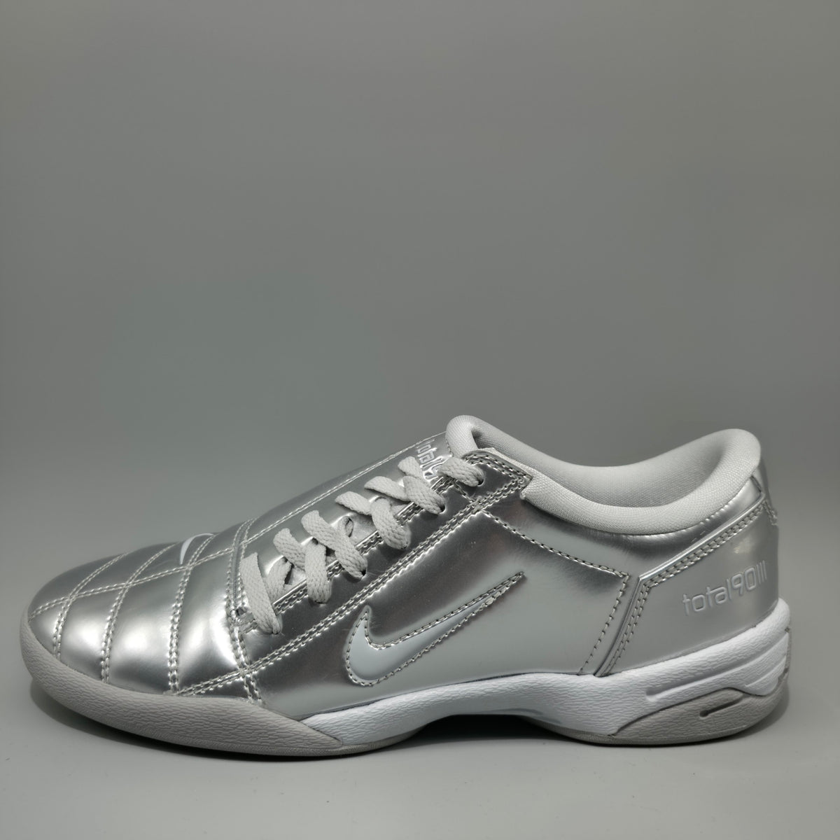 A metallic silver Nike total football shoe with white laces, a white collar, and a white sole, shown from a side profile against a gray background.