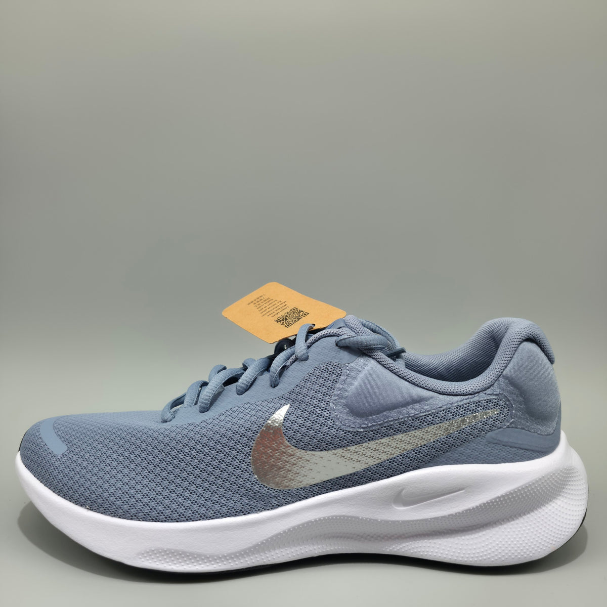 A Nike running shoe in a dusty slate blue colorway with a metallic silver Swoosh and a thick white cushioned sole. The shoe is shown with its original orange tag attached, set against a plain gray studio background.