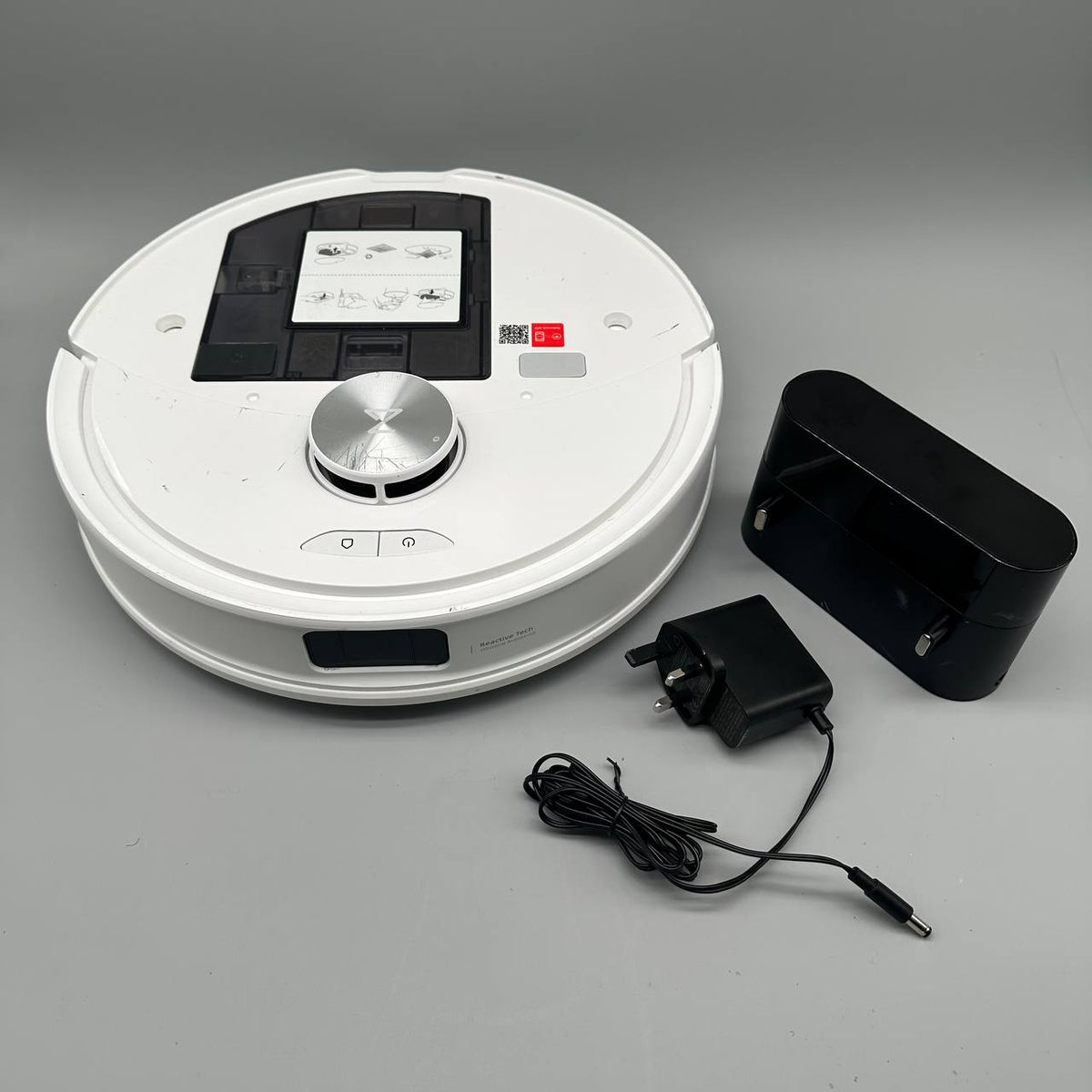A white robotic vacuum cleaner shown with its black charging dock, black power adapter, and cable on a neutral gray background.