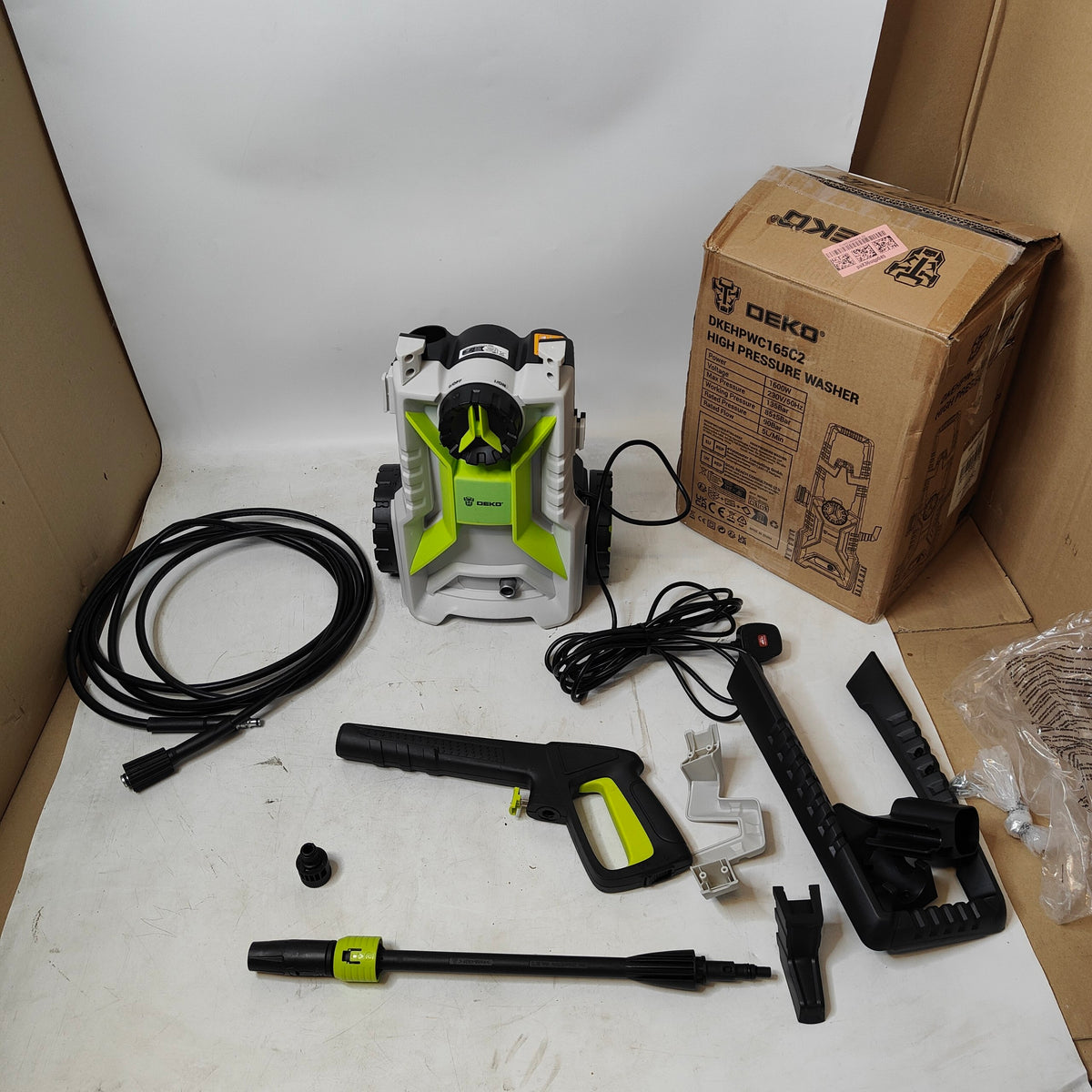 A Deko pressure washer kit laid out on a white surface, including the white and lime green pressure washer, black hose, spray wand, nozzles, mounting hardware, and a branded cardboard box.