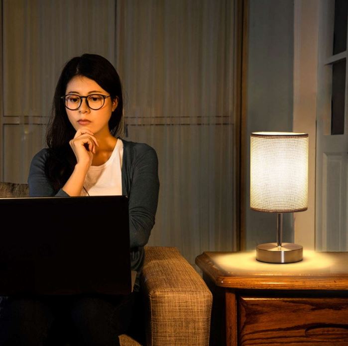 Woman sitting on a couch using a laptop in a dimly lit room with a lamp on a table next to her.